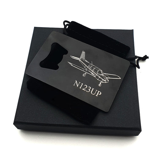 Black heavy duty bottle opener with piper pa28 aircraft and callsign engraved on the front. The bottle opener is placed on a velveteen bag and gift box.