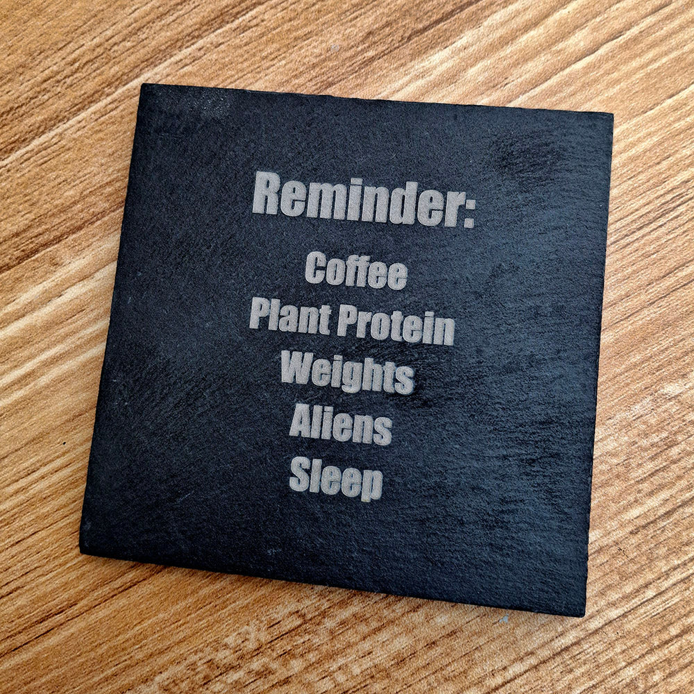 A square black slate coaster with white text reading 'Reminder: Coffee Plant Protein Weights Aliens Sleep' placed on a light brown wooden surface.