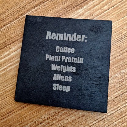 A square black slate coaster with white text reading 'Reminder: Coffee Plant Protein Weights Aliens Sleep' placed on a light brown wooden surface.