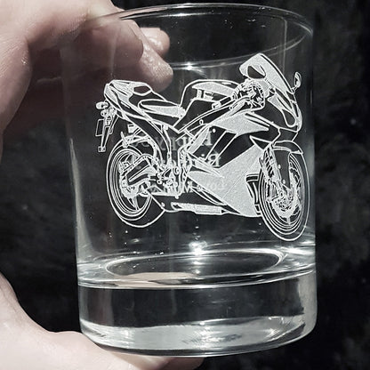 Clear glass tumbler with a motorcycle engraving held by a hand against a dark background