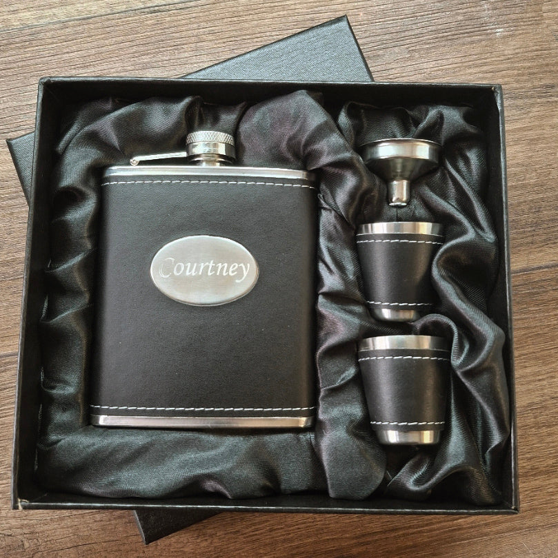Black leather flask set with 'Courtney' engraving in a box on a wooden surface