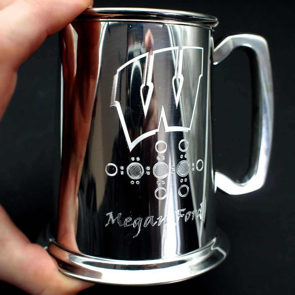 Silver mug with engraved design and text held by a hand against a black background
