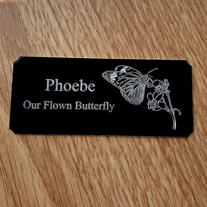 Black plaque with butterfly design and text 'Phoebe Our Flown Butterfly' on a wooden surface